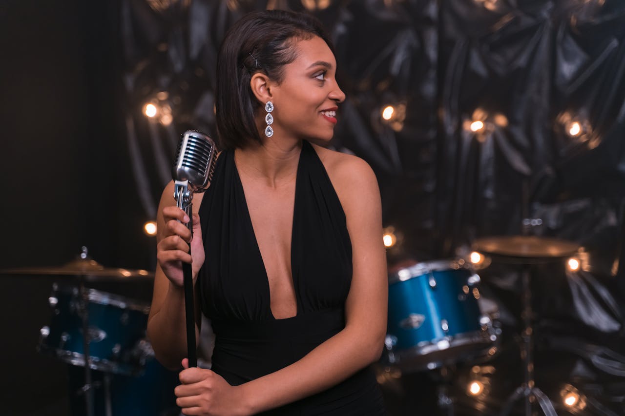 A stylish woman singing on stage with a vintage microphone and drum set.